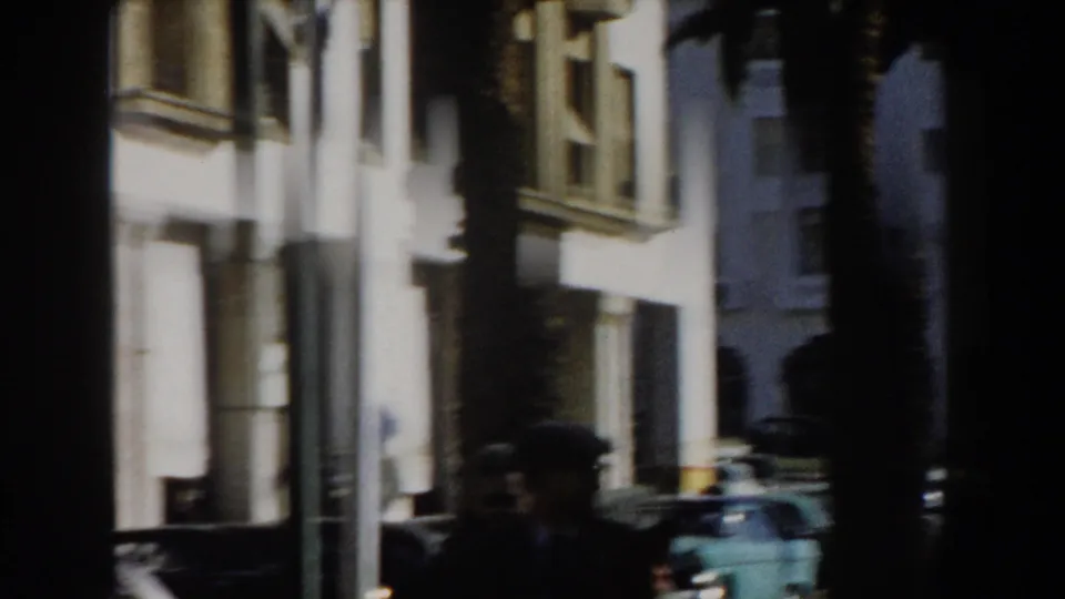 1959 Casablanca frame with palm trunks crossing white facades, a turquoise period car, and pedestrians partly obscured by shade.