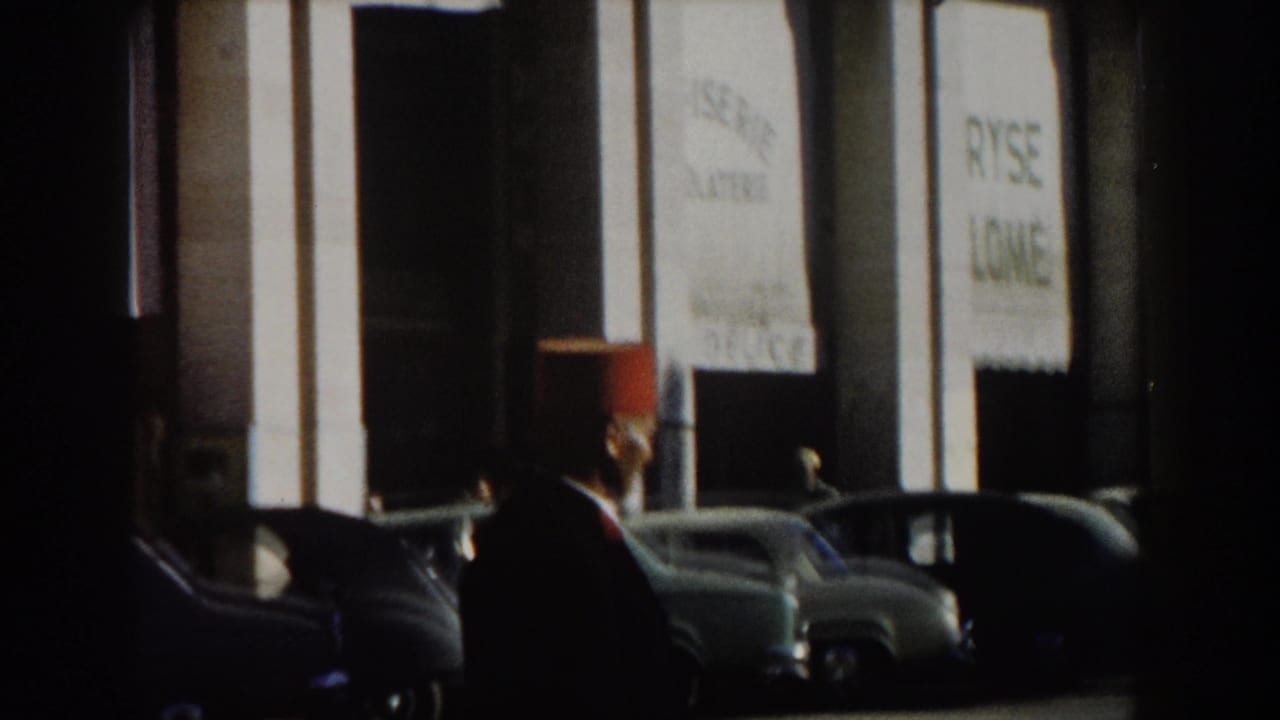 1959 Casablanca street view with a man in a red fez, grey parked cars, and a white columned facade in bright sun.