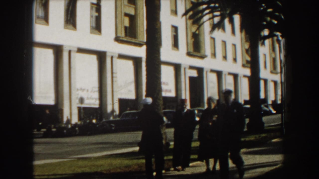 Pedestrians stand near palm trees and parked cars along a white building.