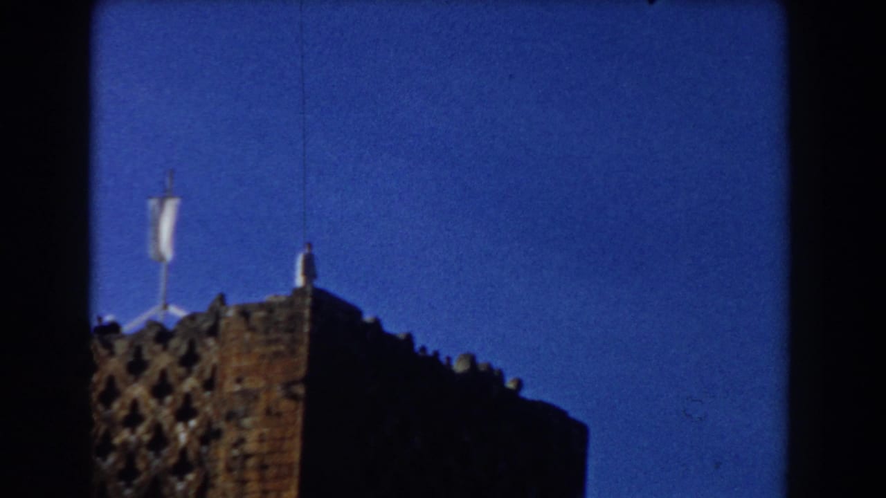 Brick building corner with a small rooftop figure, pale flag or pole, and blue sky.