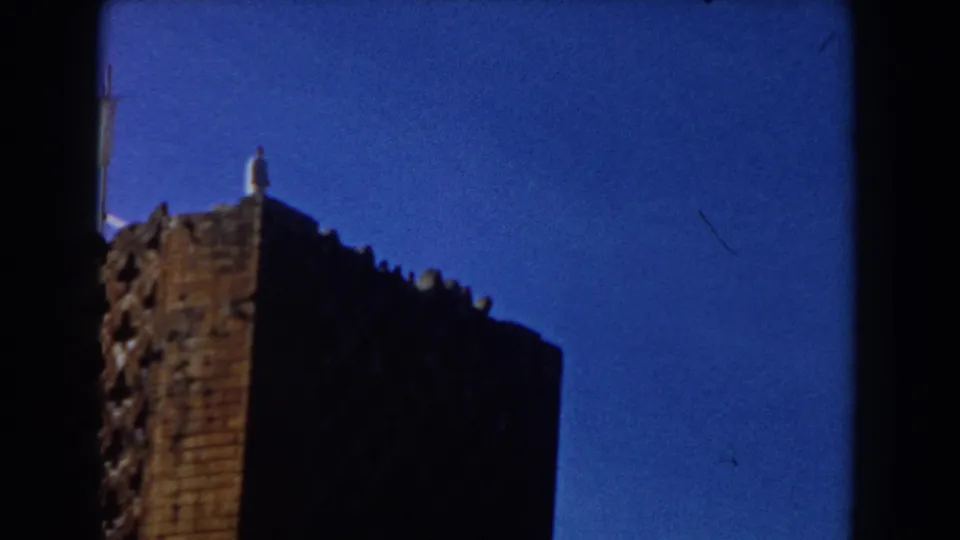 Low-angle 1959 Casablanca frame showing a weathered brick rooftop, distant standing figure, partial pale pole, and blue sky.