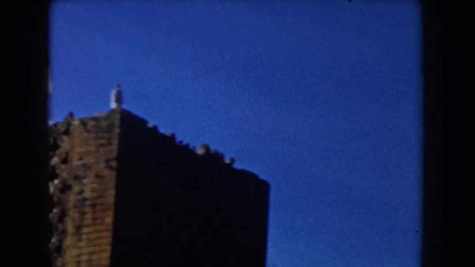 Casablanca 1959 home-movie frame with the brick building mostly in shadow and a small rooftop figure silhouetted against blue sky.