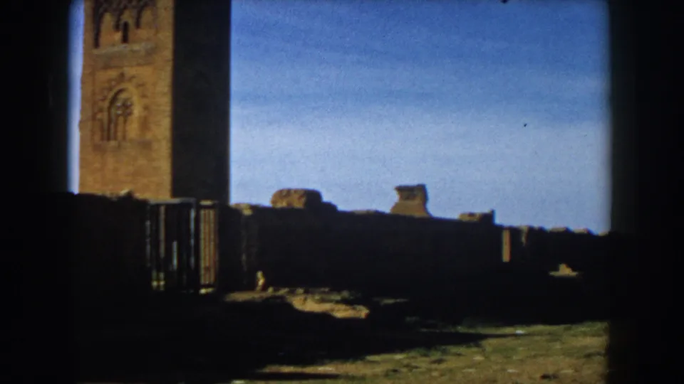 1959 Casablanca 8mm frame showing a weathered cathedral or landmark facade through deep foreground shadow with low walls and blue sky.