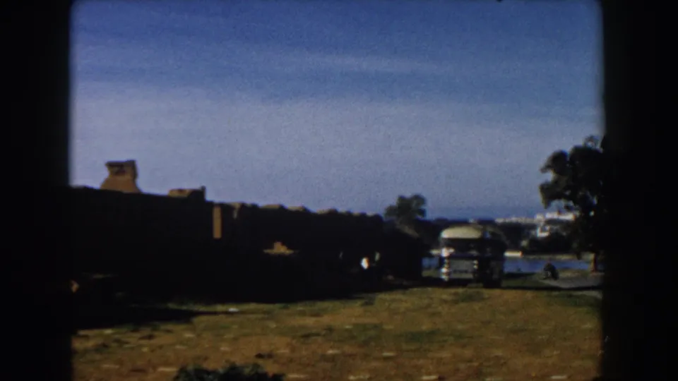 1959 Casablanca frame showing a bus beside stone ruins, with trees and distant city buildings in the background.