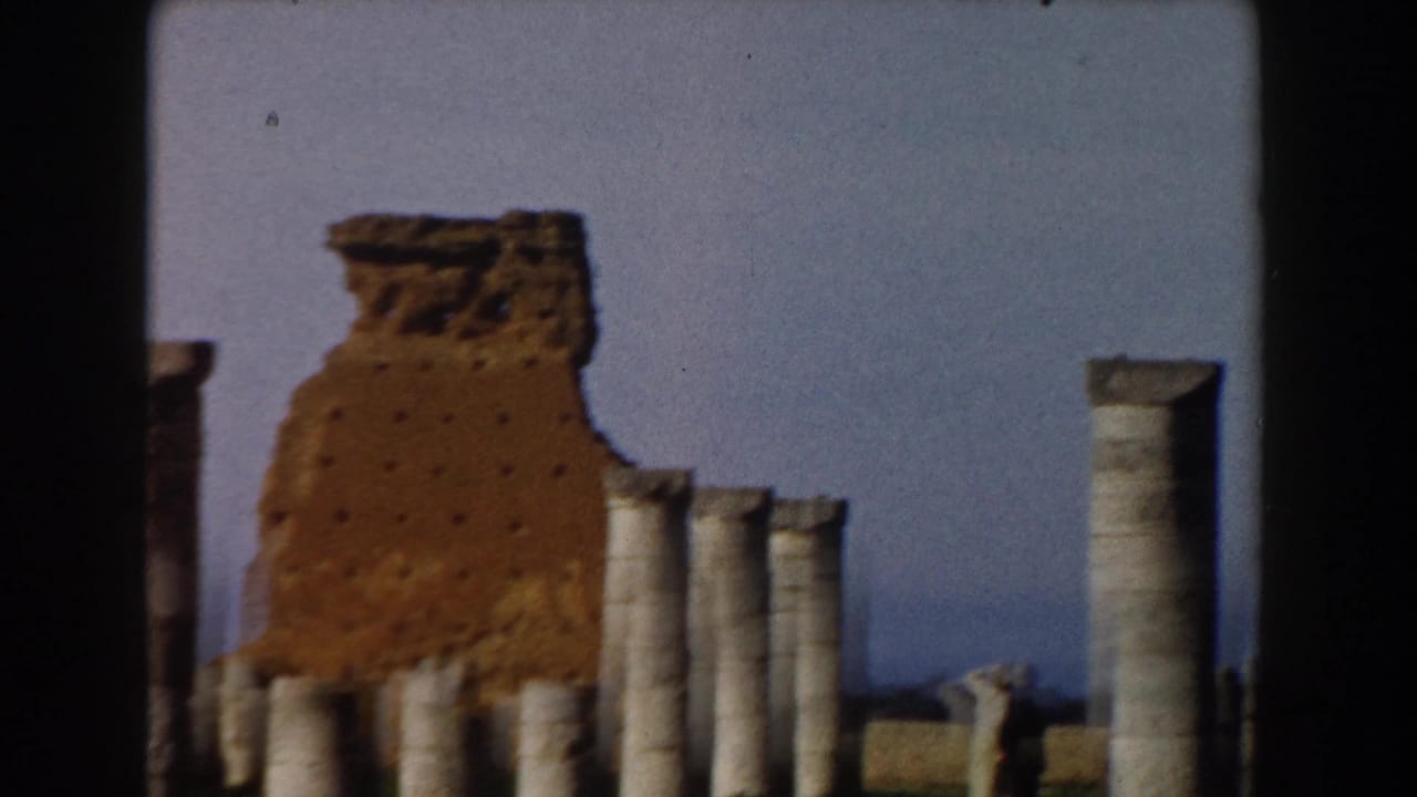 Crumbling brick wall and rows of stone columns under a blue sky.