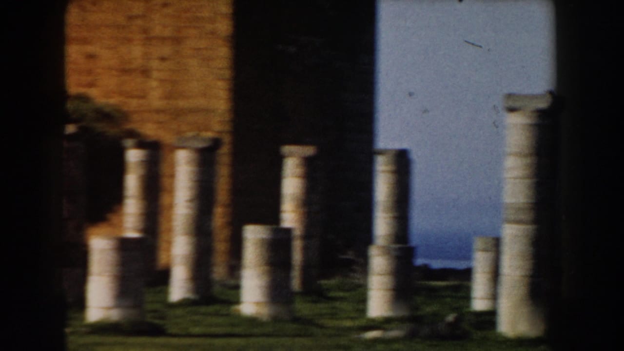 Stone columns and column fragments standing on grass near a masonry wall.