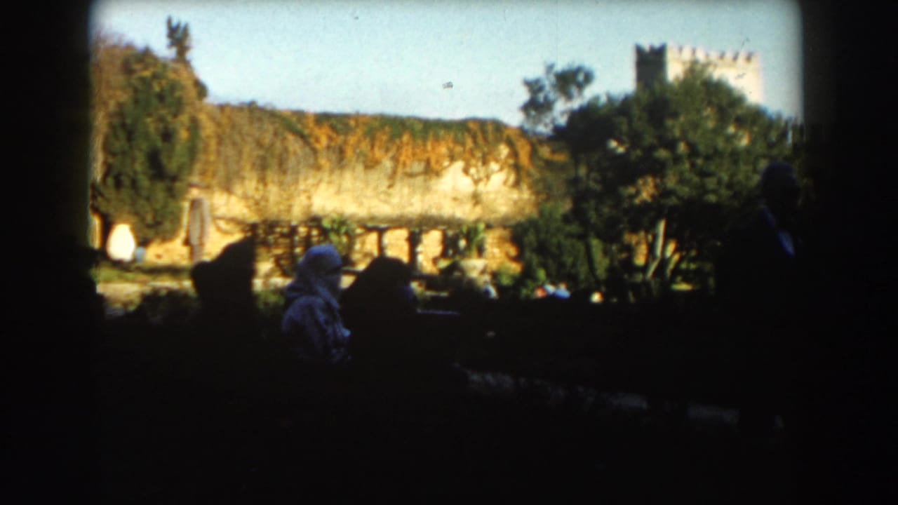 1959 Casablanca home-movie frame with shaded seated figures, a bright stone wall, trees, garden planting, and a tower in the background.