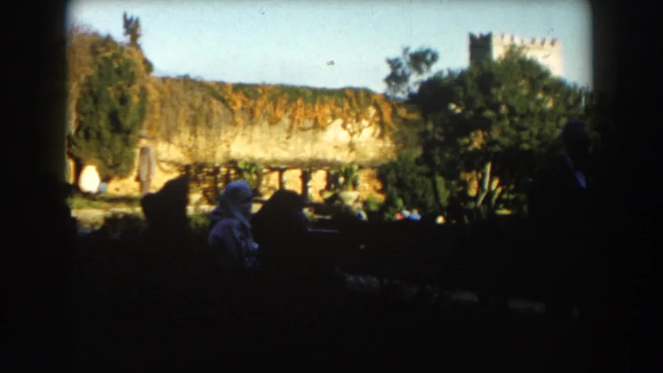 1959 Casablanca home-movie frame with shaded seated figures, a bright stone wall, trees, garden planting, and a tower in the background.