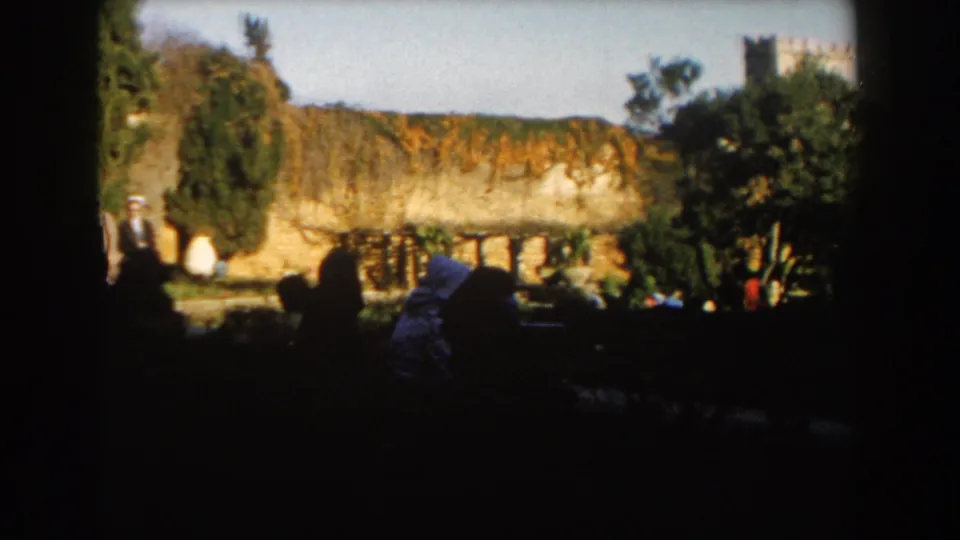 1959 Casablanca outdoor frame with a man in a hat at left, seated silhouettes in shade, a sunlit wall, trees, and garden plants.