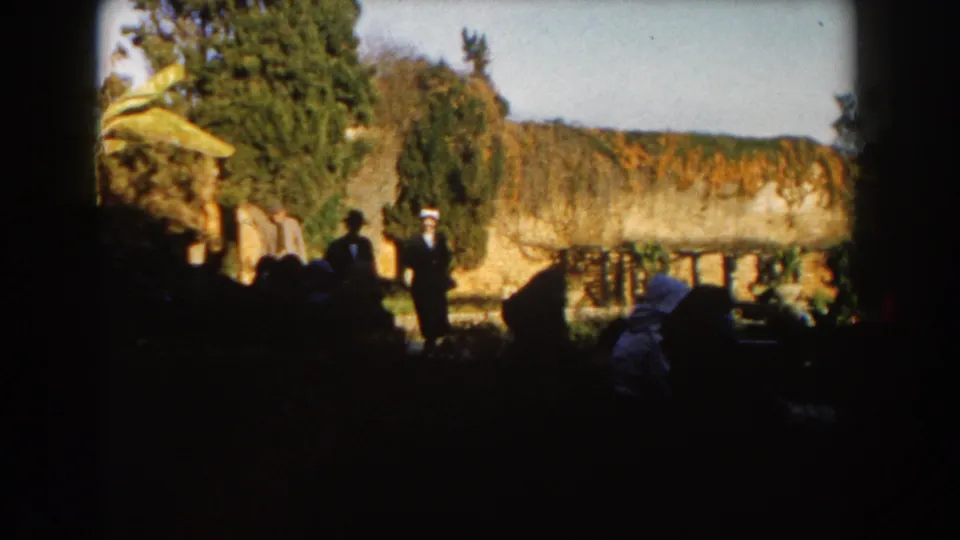 1959 Casablanca home-movie frame with suited visitors in shade, leafy trees, garden planting, a bright stone wall, and sky beyond.