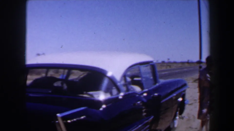 1958 Las Vegas-area home-movie frame showing a classic car parked near a dusty desert roadside and a partially visible adult figure.