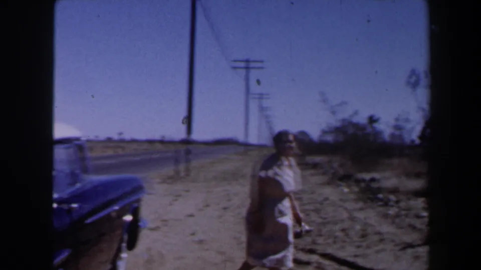 1958 Las Vegas-area frame of a woman in a light dress and hat standing by a desert roadside with utility poles in the distance.
