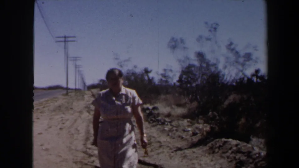 The woman is closer in frame as she walks on a desert roadside near Las Vegas, with brush and utility lines behind her.