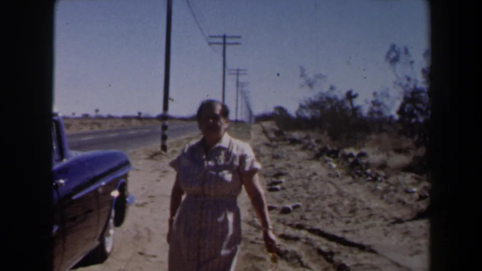 A woman near the camera stands or walks beside a dark car at the edge of a dusty Las Vegas-area desert road.