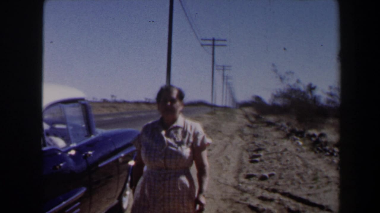 Woman walking beside a vintage car on a dusty roadside with utility poles behind her.