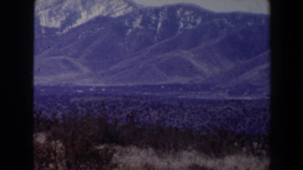 Likely Las Vegas-area 1958 home-movie frame of snow-streaked mountains beyond sparse desert vegetation.