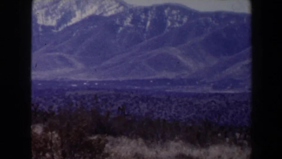 Likely Las Vegas-area 1958 home-movie frame of snow-streaked mountains beyond sparse desert vegetation.