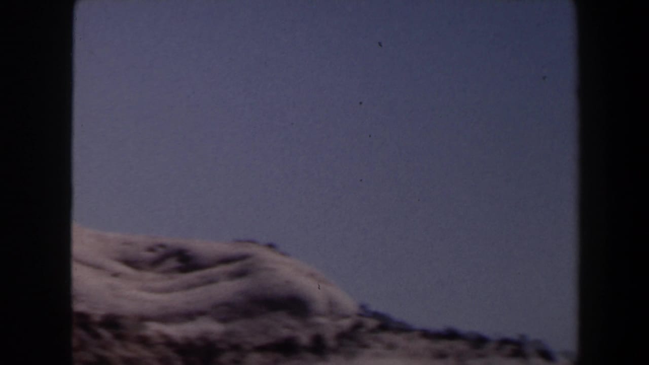 Final 1958 Las Vegas-area frame with a low sandy ridge, sparse scrub, and broad blue sky from a moving home-movie shot.