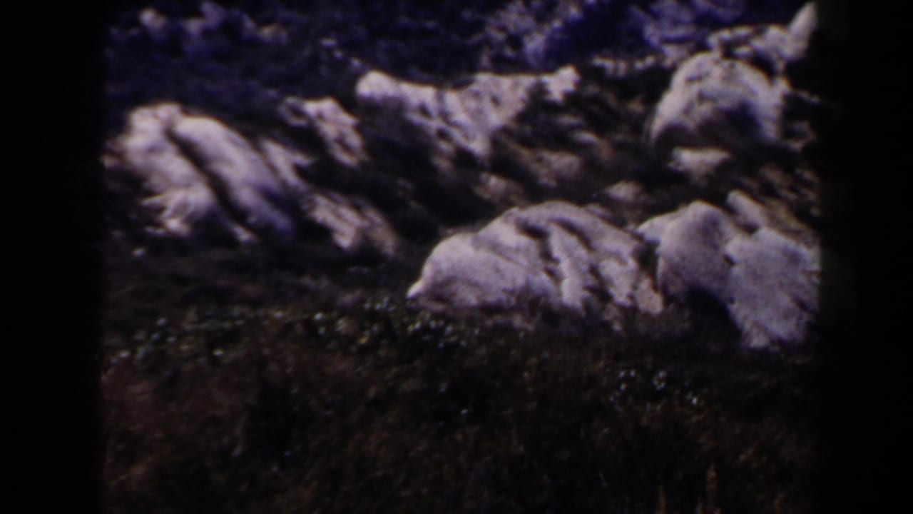 1958 Las Vegas-area 8mm frame focused on pale weathered rock formations and sparse dark vegetation under a purple-blue color cast.