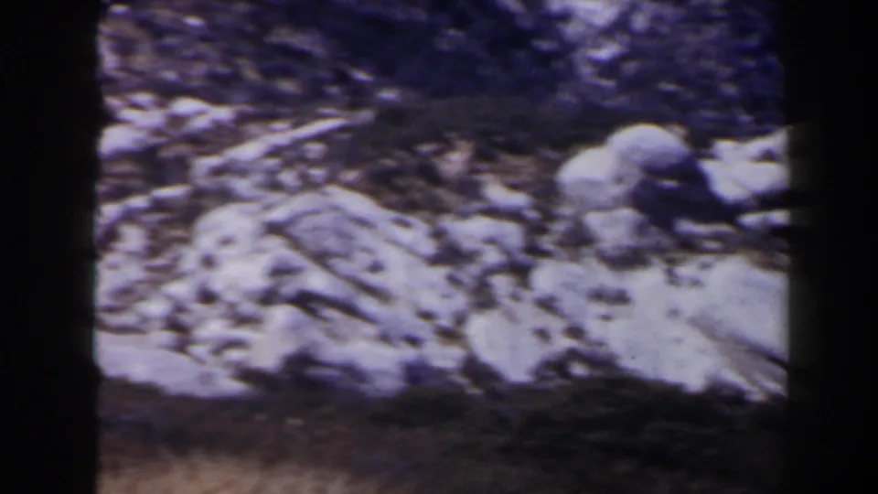 1958 8mm frame of a rocky mountain slope near Las Vegas, with scattered melting snow and a purple-blue aged-film cast.