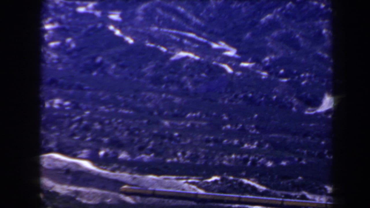 1958 8mm frame showing a blue-tinted snow-covered mountain slope above a darker lower landscape, with visible film edges.