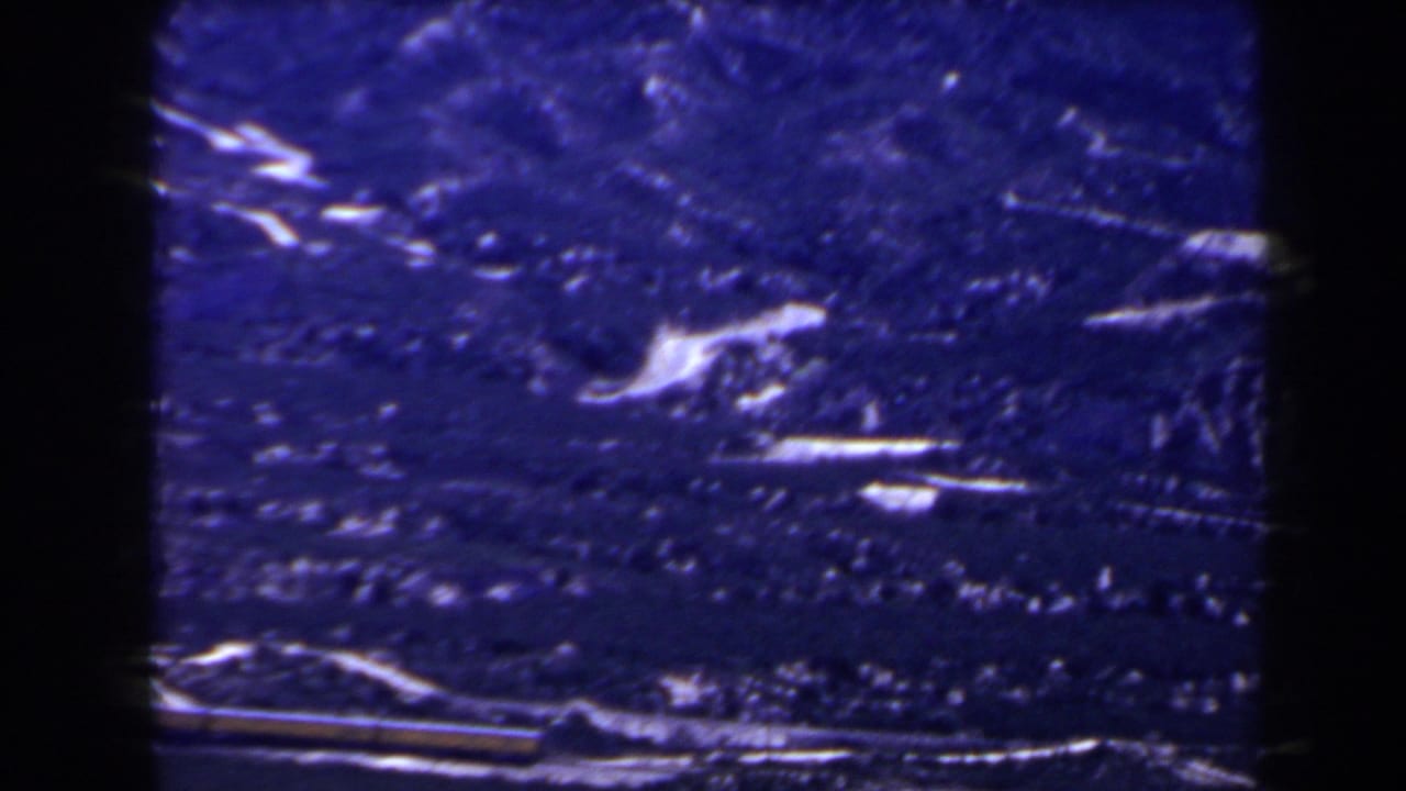 Blue-shifted 1958 8mm view of snowy mountain terrain and darker valley ground, with a possible rail or train line near the bottom.
