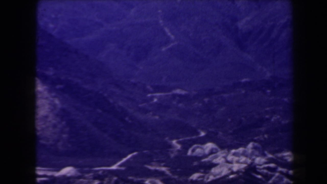 1958 Las Vegas-area 8mm frame showing shadowed valley ground, faint pale paths, and rocky foreground details under a purple cast.