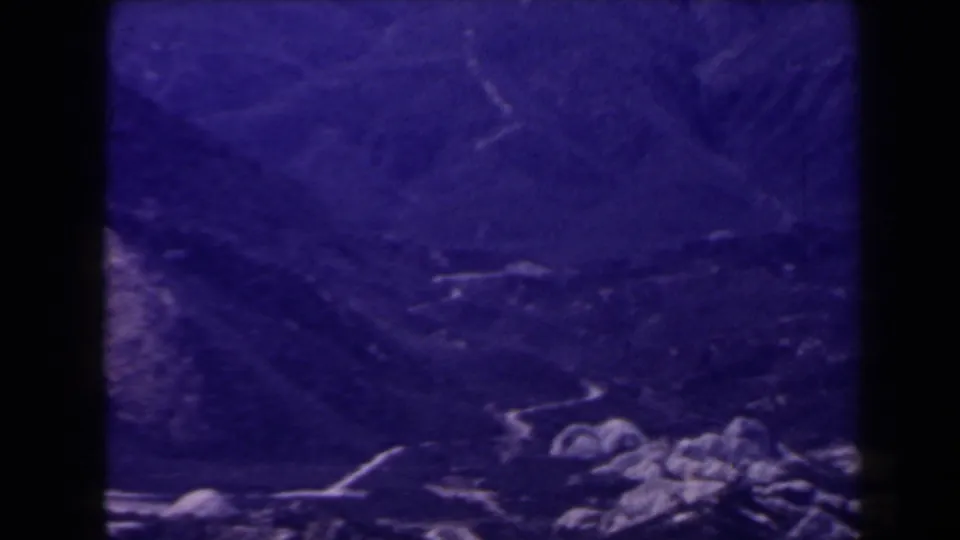 1958 Las Vegas-area 8mm frame showing shadowed valley ground, faint pale paths, and rocky foreground details under a purple cast.