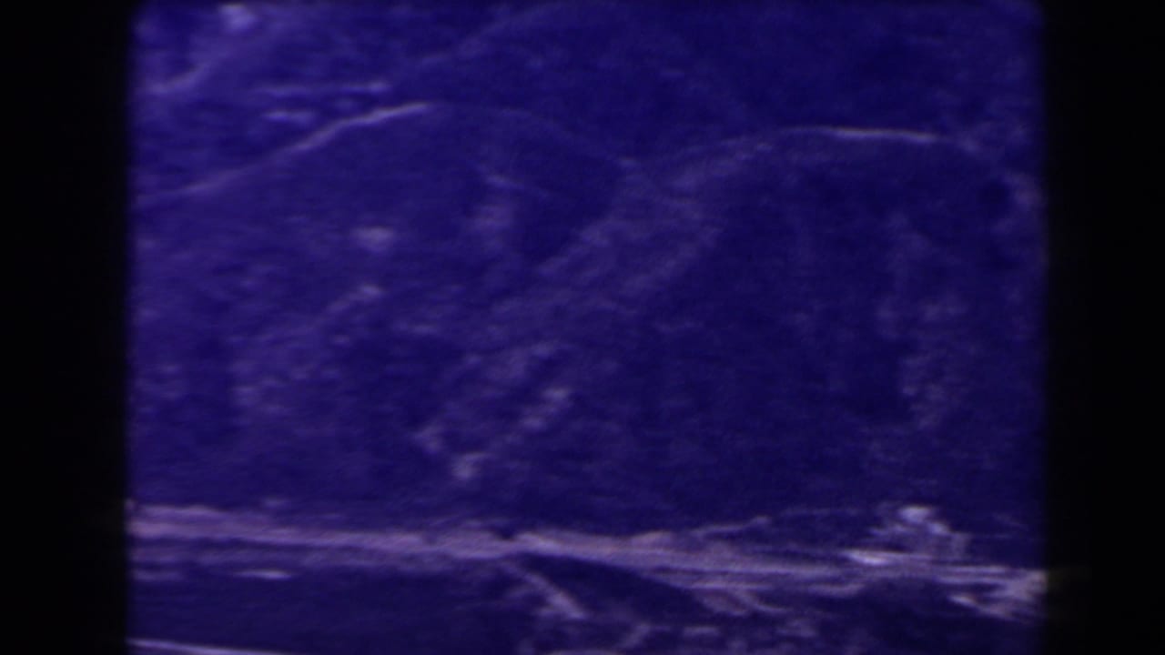 1958 Las Vegas-area 8mm frame with a purple mountain valley, faint path traces, and a pale rocky strip along the lower edge.