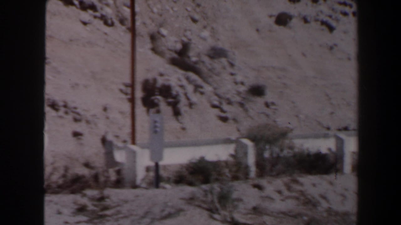 Las Vegas-area desert roadside view with a pale concrete barrier, vertical pole, scrub, and rocky slope in 1958 home-movie footage.