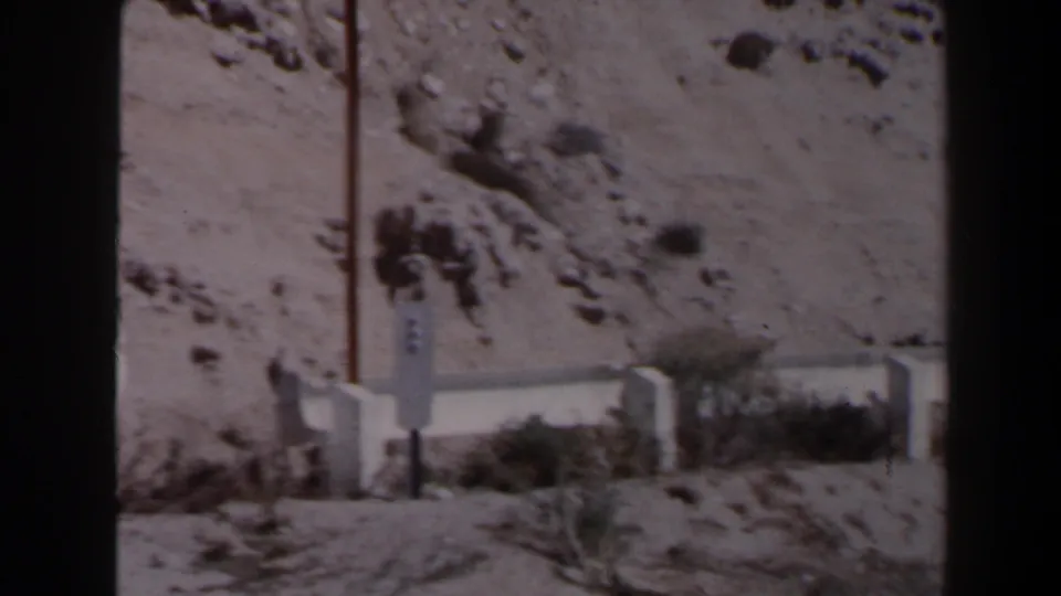 Las Vegas-area desert roadside view with a pale concrete barrier, vertical pole, scrub, and rocky slope in 1958 home-movie footage.