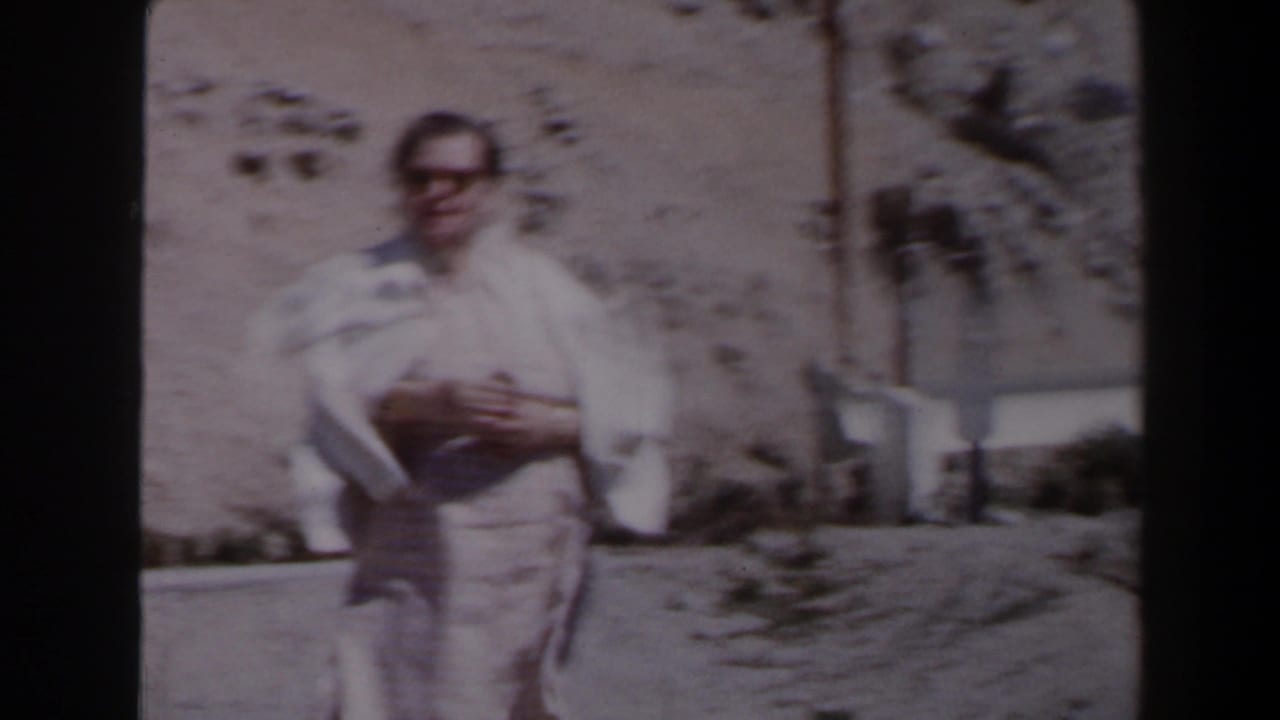 A man in light clothing faces the camera on a Las Vegas-area desert road, with a barrier and rocky hillside behind him.