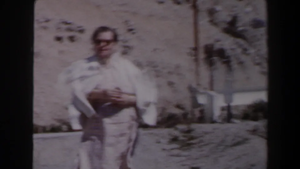 A man in light clothing faces the camera on a Las Vegas-area desert road, with a barrier and rocky hillside behind him.