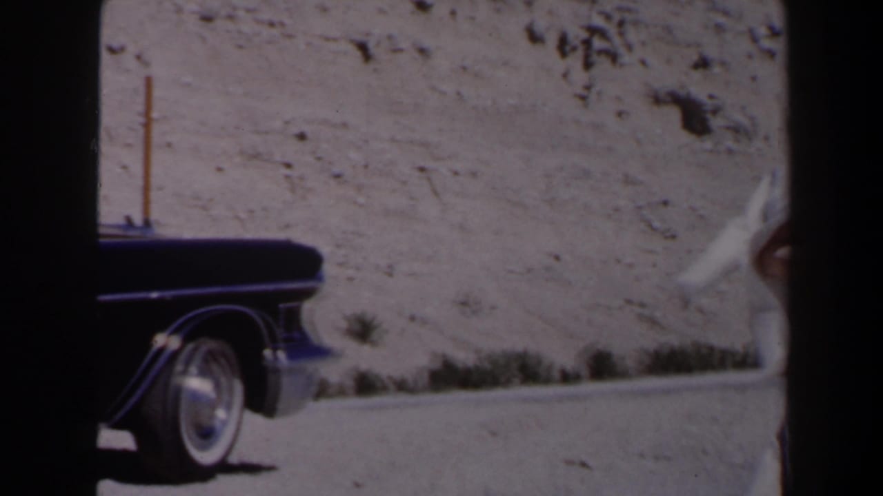 Dark mid-century car stopped on a sunlit Las Vegas-area desert road, with a pole, scrub, and rocky hillside nearby.
