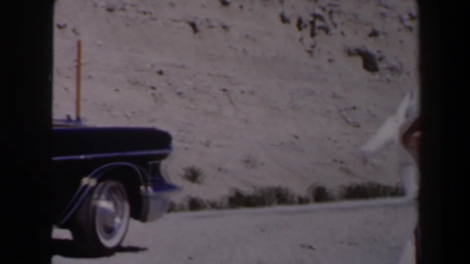 Dark mid-century car stopped on a sunlit Las Vegas-area desert road, with a pole, scrub, and rocky hillside nearby.