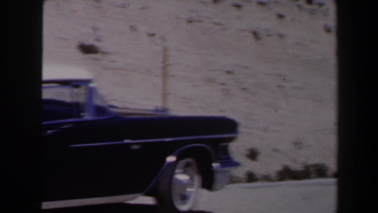 Dark 1950s Cadillac passing a rocky desert roadside.