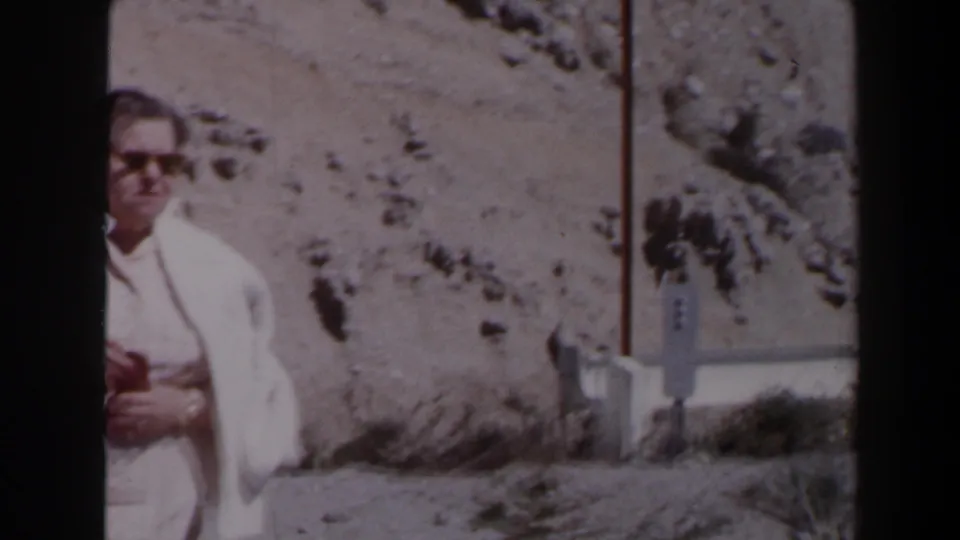 A man in a light shirt and sunglasses holds a camera near a rocky Las Vegas roadside in 1958 home-movie footage.