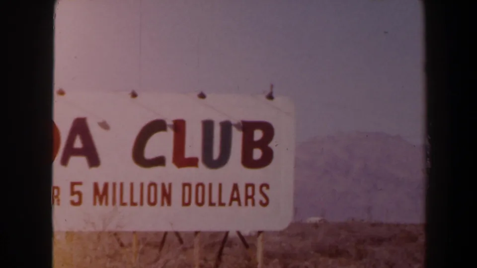 1958 Las Vegas-area 8mm frame showing a cropped white roadside sign with red CLUB lettering, partial 5 MILLION DOLLARS text, dry brush, and distant mountains.