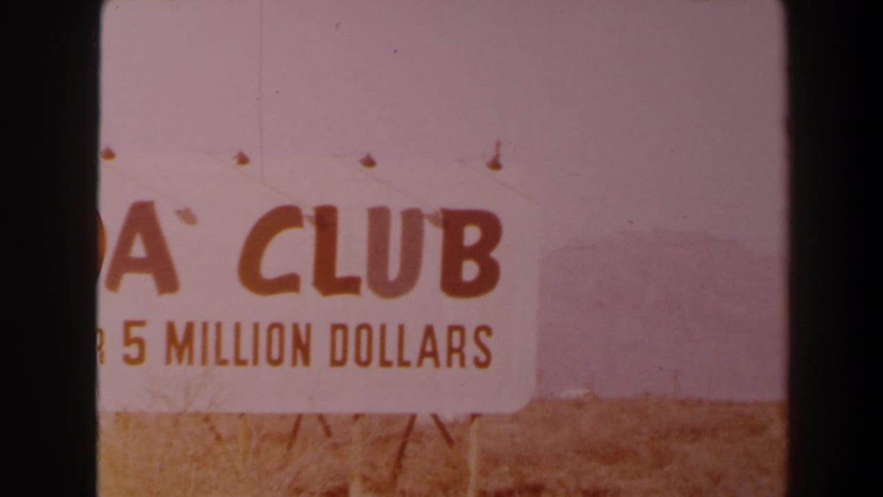 1958 Las Vegas-area frame with a roadside sign showing CLUB and 5 MILLION DOLLARS, set before low desert mountains under hazy light.