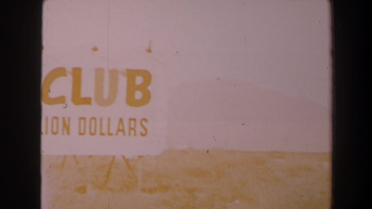 Faded 1958 8mm frame with the roadside sign partly cropped, showing CLUB and partial DOLLARS text over pale desert terrain.