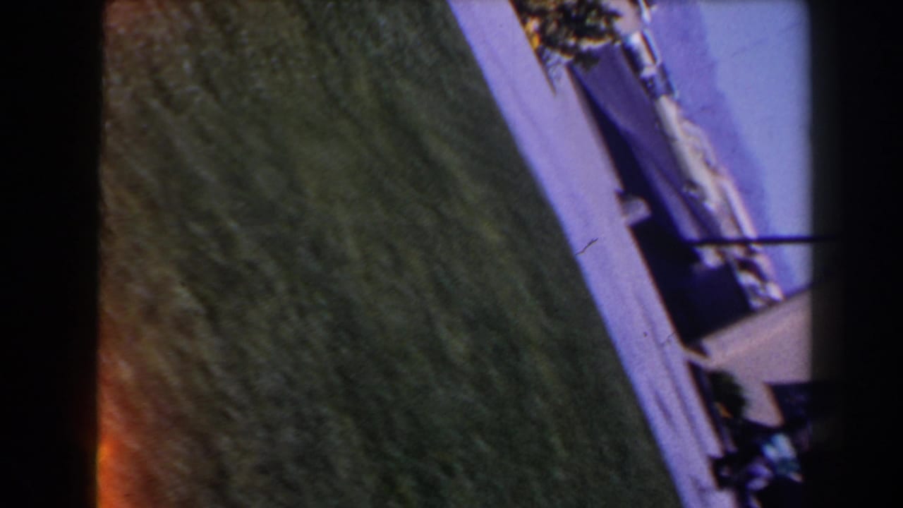Tilted 1958 Las Vegas exterior view with grass, pavement, and a partial building edge seen through grainy 8mm film.