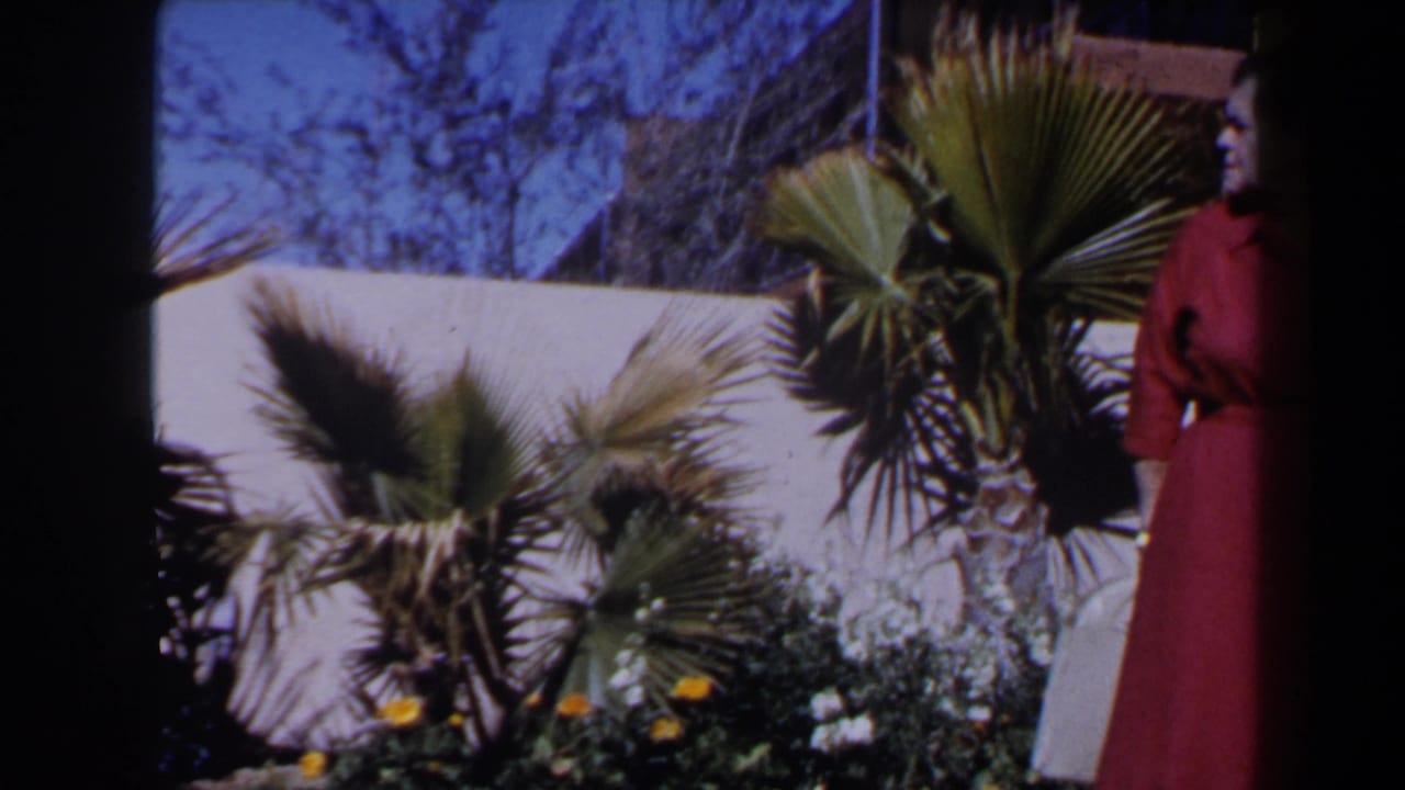 1958 Las Vegas frame with a mature woman in a red dress standing beside a palm-filled garden and white wall.