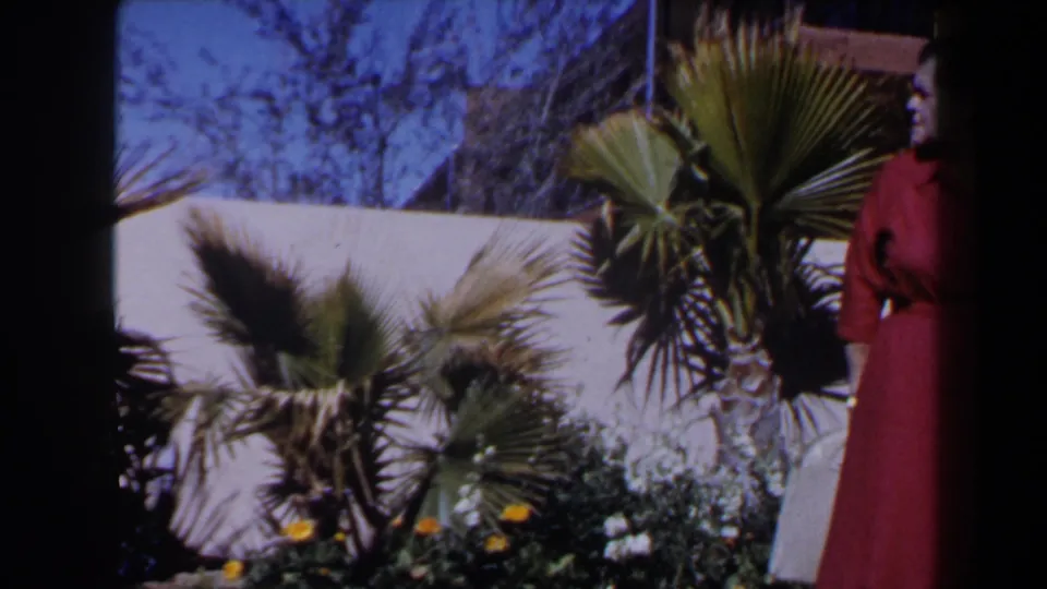 1958 Las Vegas frame with a mature woman in a red dress standing beside a palm-filled garden and white wall.