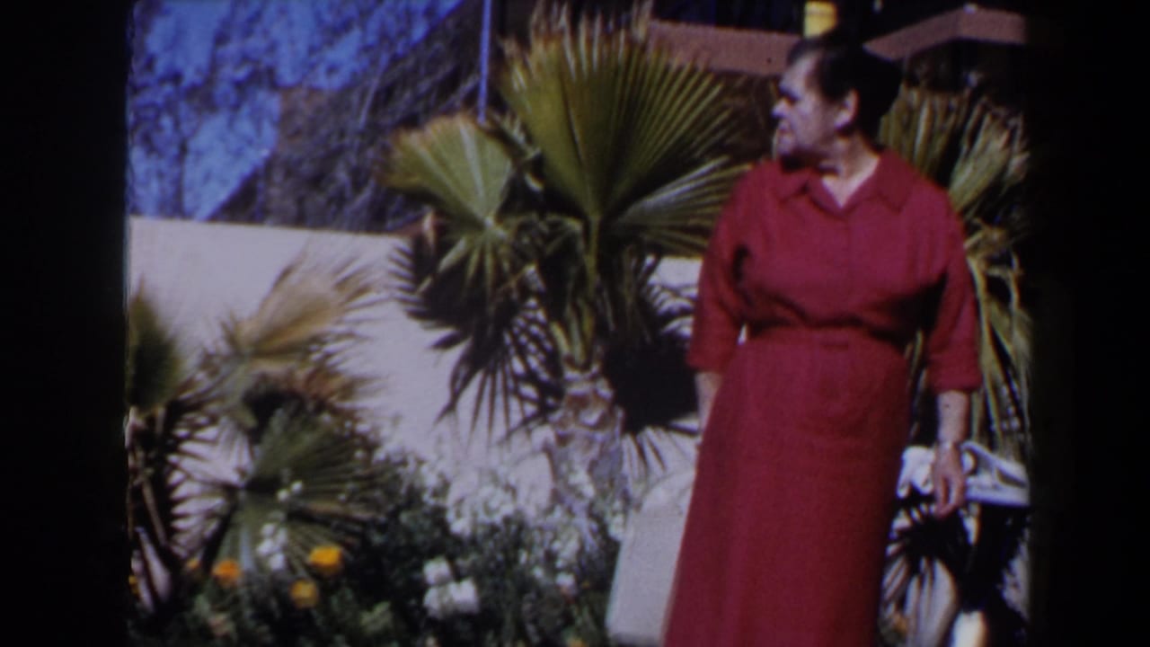 Mature woman in a red dress looks to the side in a 1958 Las Vegas garden with palms, flowers, and a white wall.