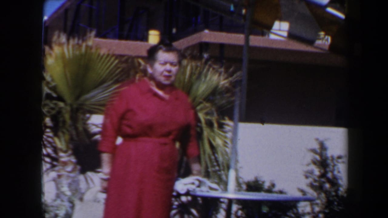 Woman in a red dress stands near palm plants outside a low building.