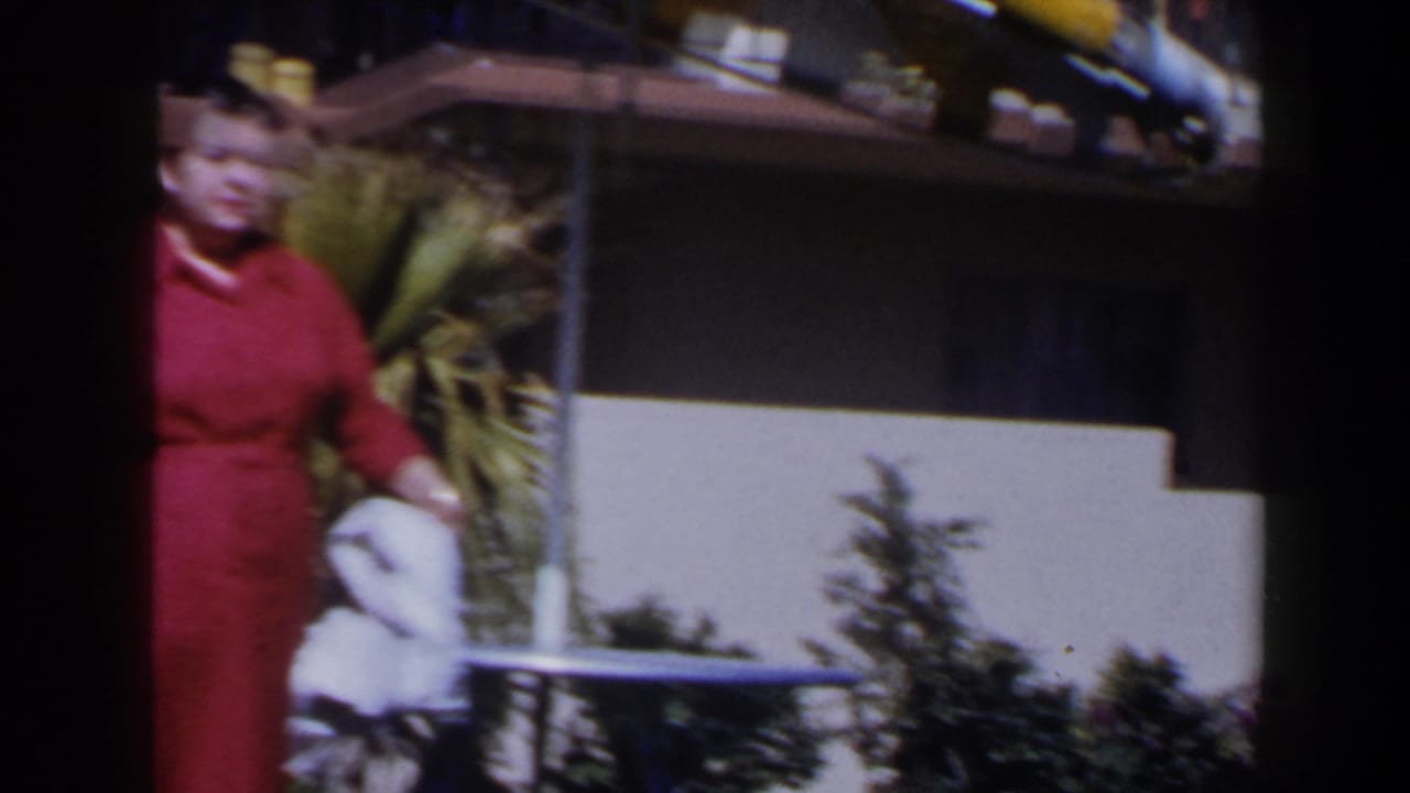 The woman in the red dress remains near a small outdoor table and palm plants, with balcony railings and a sunlit building behind her.
