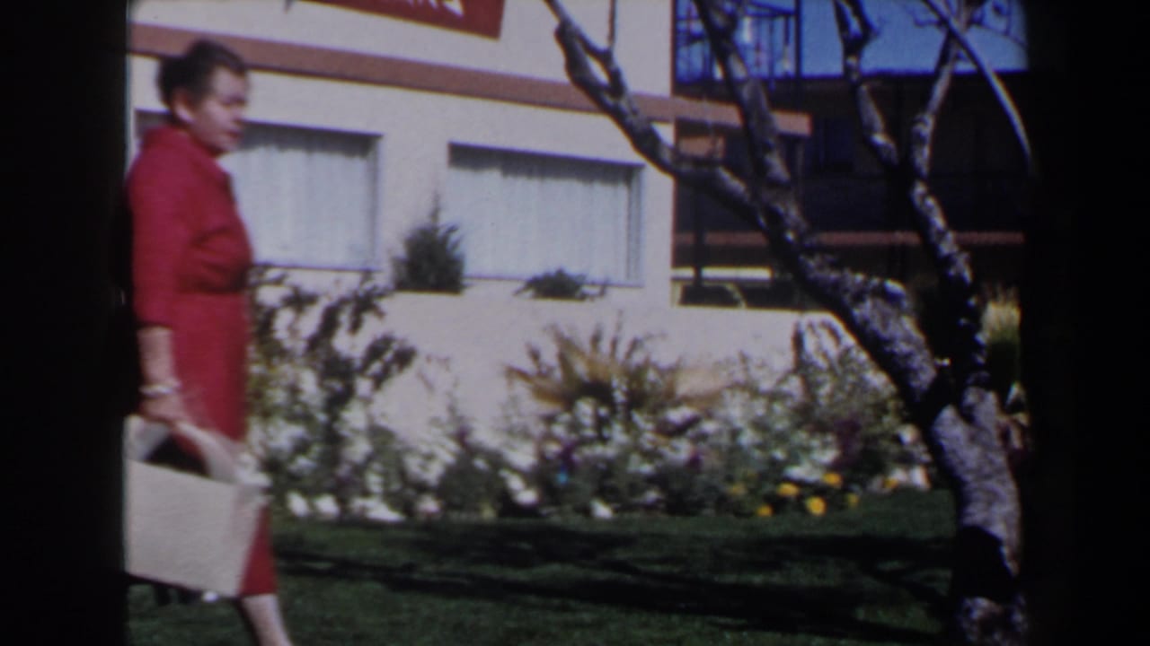A woman in a red dress walks through a Las Vegas landscaped area in 1958, carrying a light-colored bag near trees and windows.
