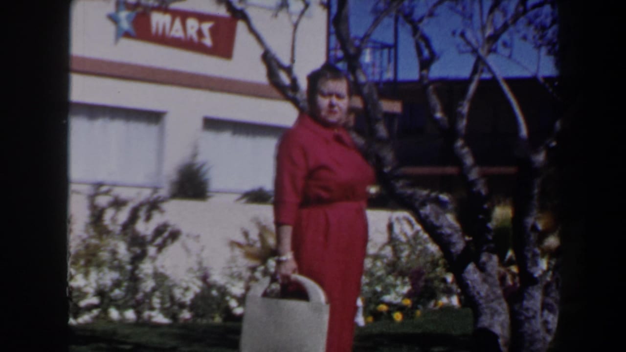 A woman in red stands or turns toward the camera in a 1958 Las Vegas garden setting, with bags, trees, windows, and a partial MARS sign behind her.
