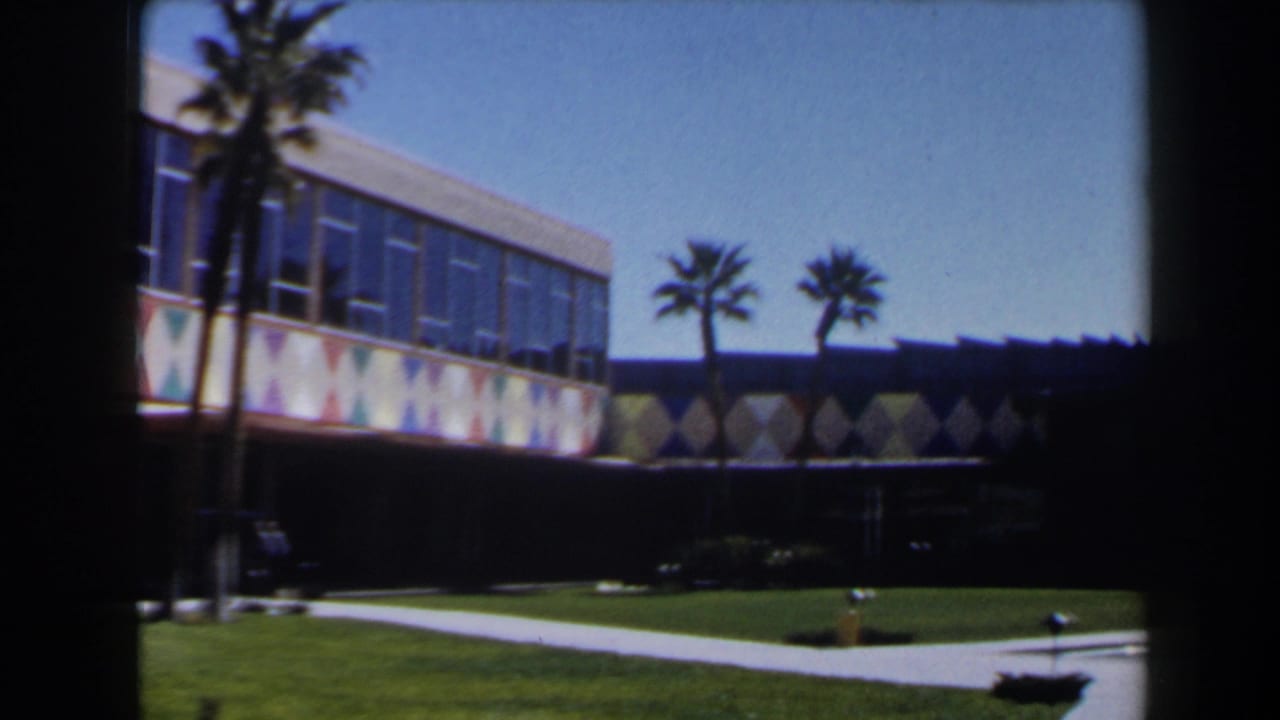 Las Vegas 1958 home-movie frame showing a palm-lined modern exterior with lawn, sidewalk, and colorful geometric wall decoration.