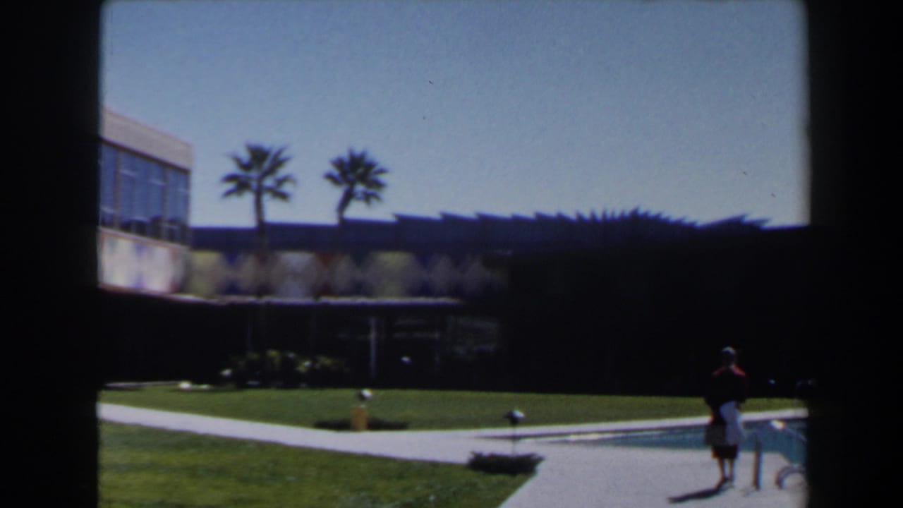 Las Vegas 1958 exterior with palm trees, lawn, shaded building, pool edge, and a small distant figure near the water.